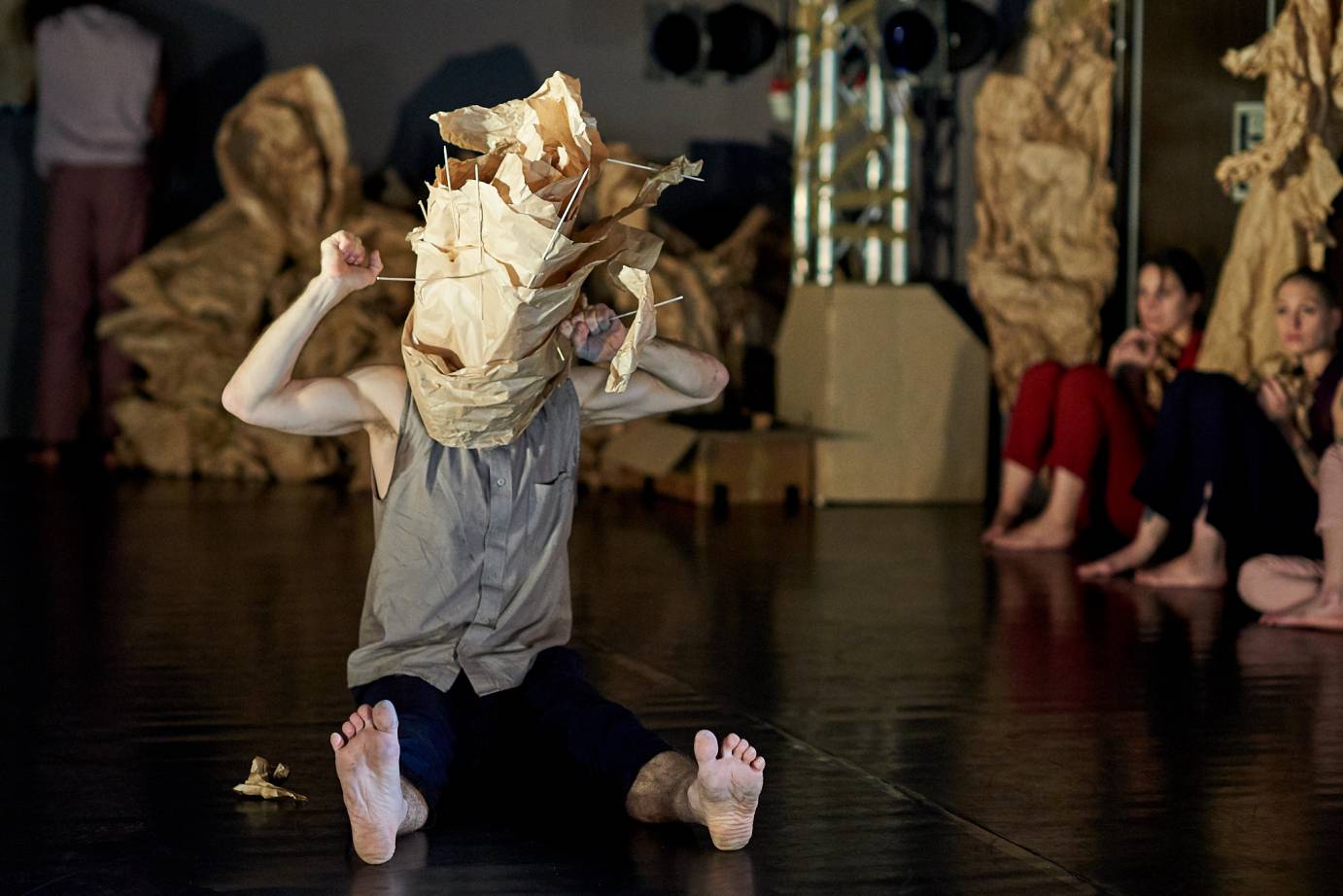 A man whose head is covered by a weird paper sculpture sits with his feet flexed